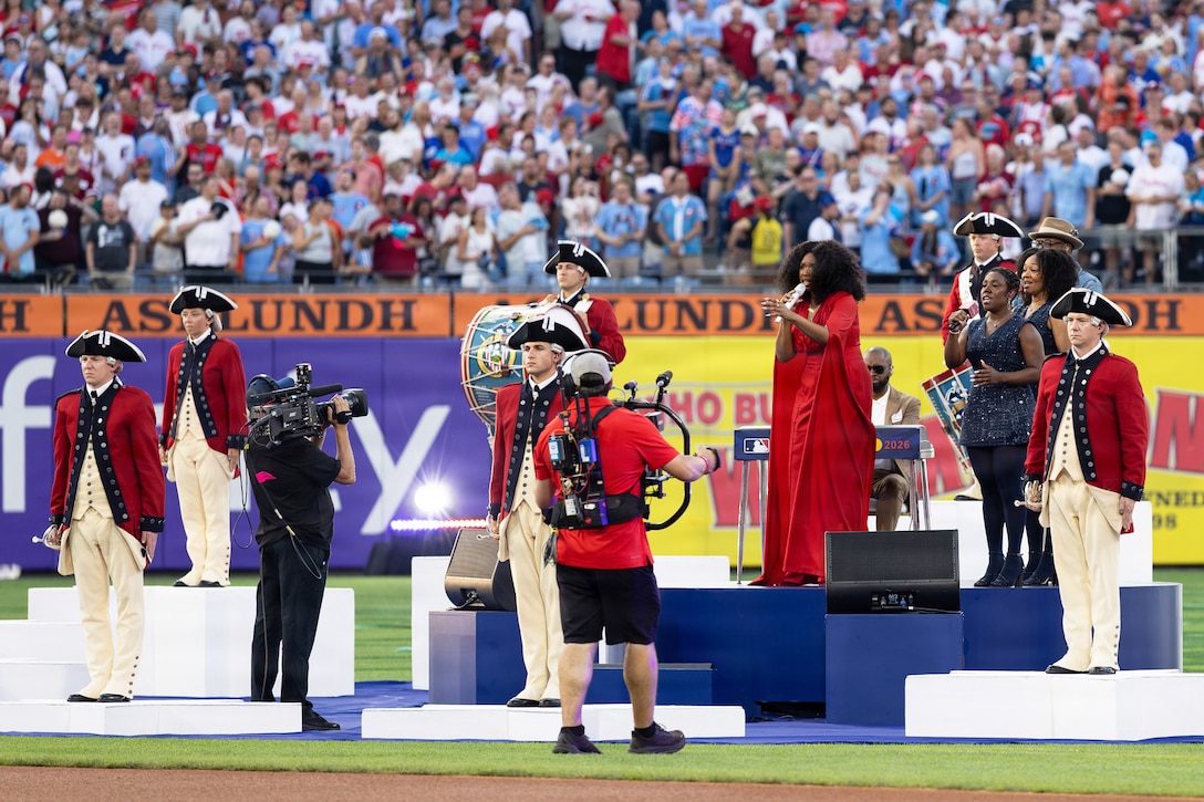 Soldiers in ceremonial uniforms stand on white blocks surrounding a singer performing on a blue stage on a baseball field in front of dozens of spectators.