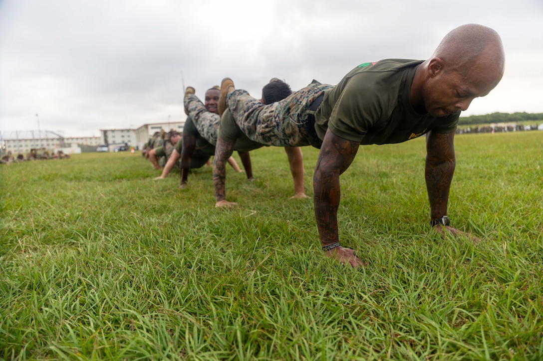 Marines rest their legs on fellow Marines' shoulders to perform squad pushups in the grass.