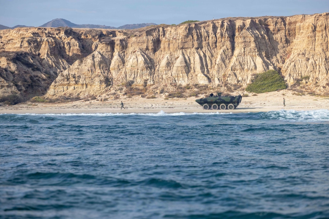 A Marine runs in front of an amphibious combat vehicle as another Marine follows it on the sand in front of a cliff with the ocean in the foreground.