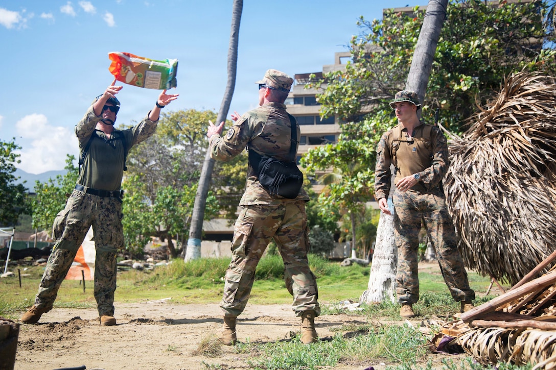 A soldier, a Marine and a sailor pass a bag in an assembly line near trees during the day.