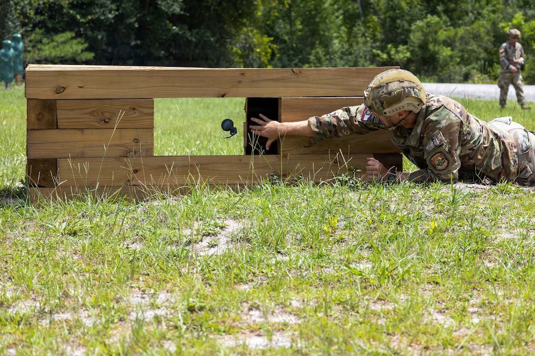 A soldier tosses a simulated grenade through the window of a wooden structure while lying in the grass.