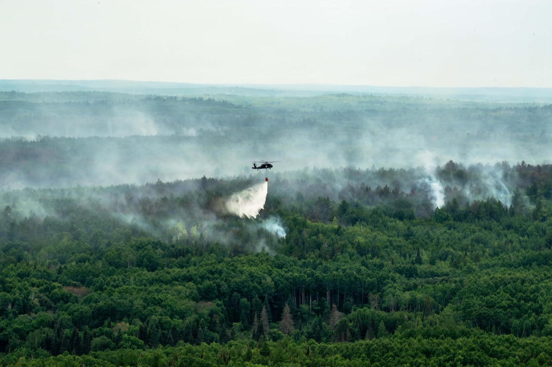 A wide view of a helicopter in a hazy sky dumping water from a bucket onto a smoky forest.