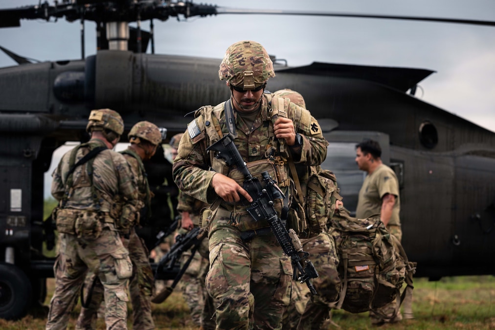 A soldier in a camouflage uniform carrying a backpack and weapon walks in front of other soldiers gathered in front of a black helicopter under a gray sky.
