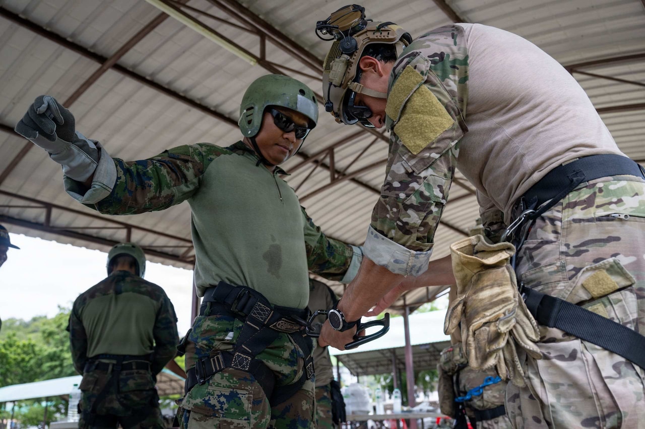A man wearing a military camouflage uniform holds his arms up while another man in similar attire does a safety check in the first man’s rappelling gear.