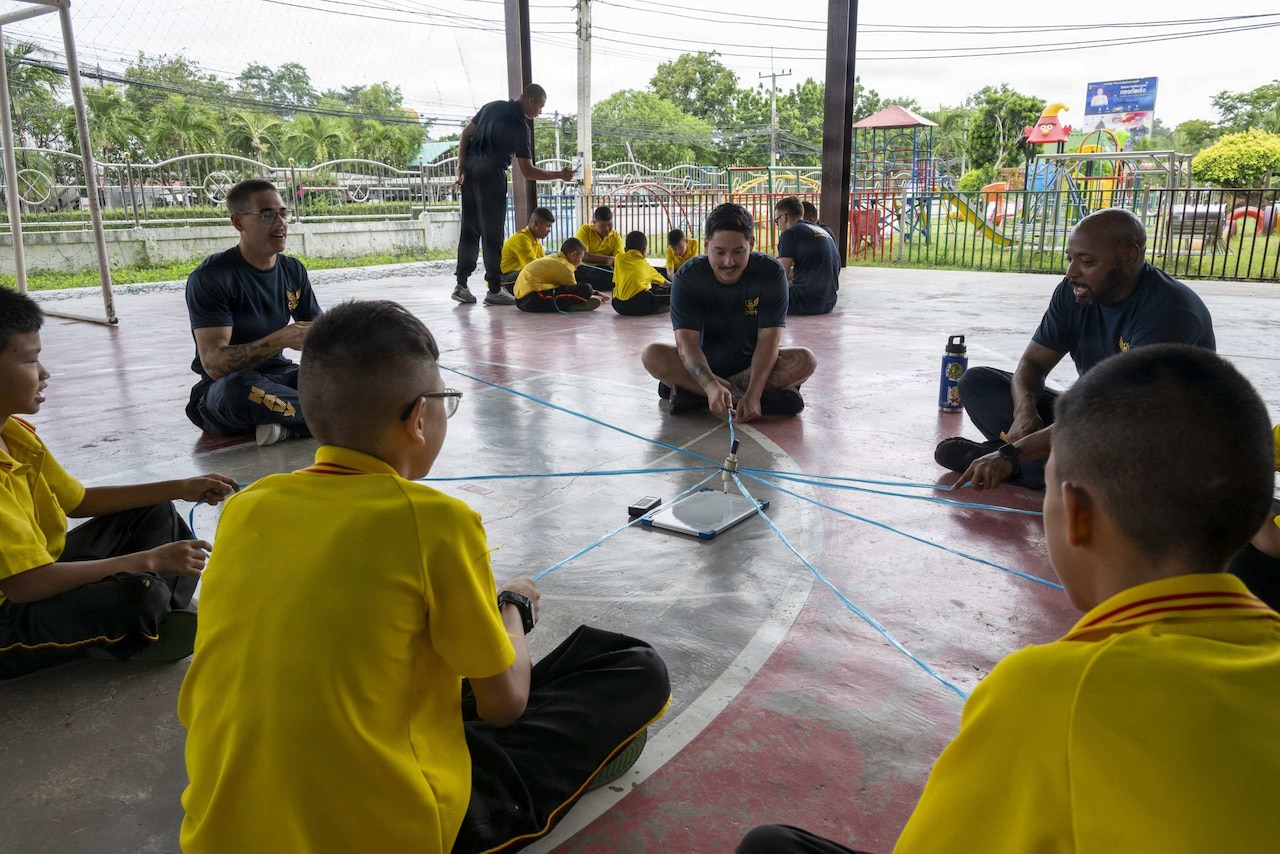 Three men sit in a circle with children playing a game. There is a similar group in the background.