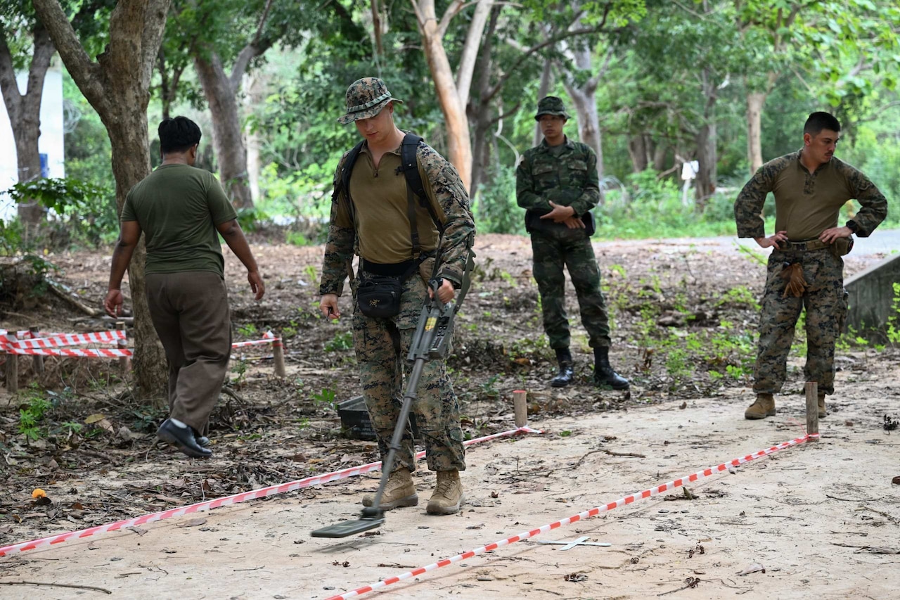 A man dressed in a military camouflage uniform holds mine sweeping equipment over the sand in a marked lane as two other men wearing similar attire watch and another man walks by.