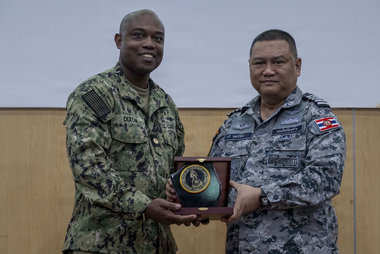 Two men wearing military camouflage uniforms hold a wooden box and an award while posing for a photo.