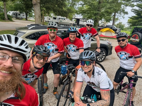Seven cyclists in cycling gear pose for a photo with their bicycles at a campground.