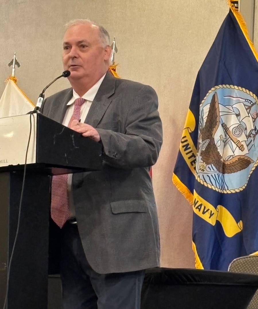 Man in suit speak at podium in front of flags.