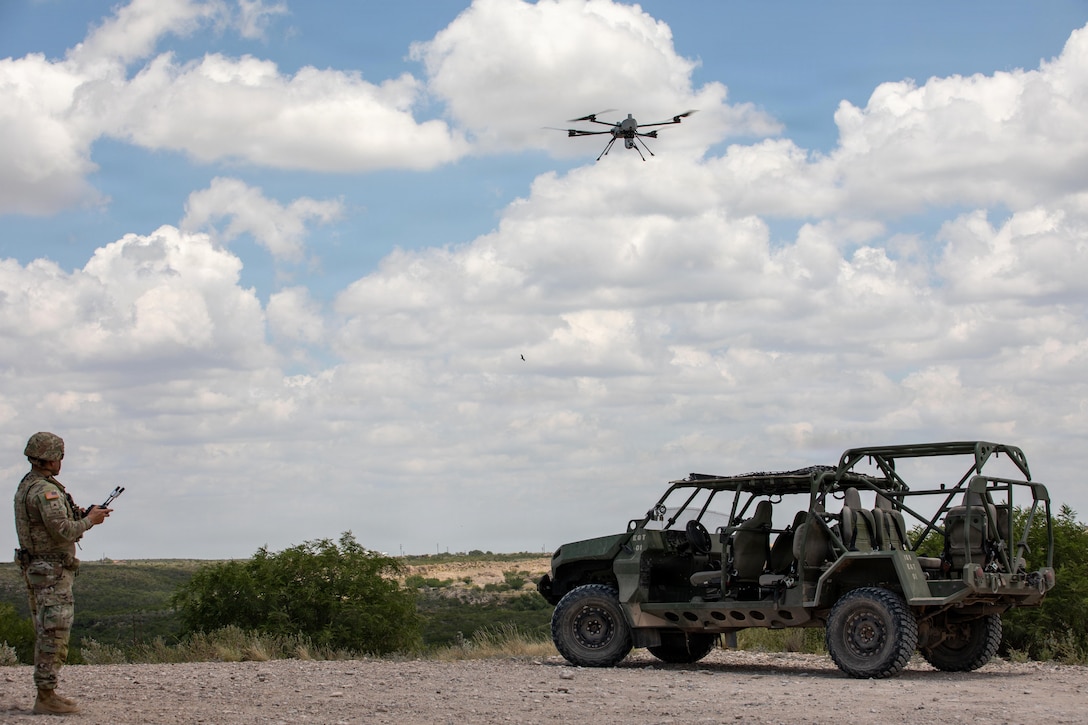 A soldier holds a control device as a drone hovers above a vehicle parked nearby on dirt terrain.