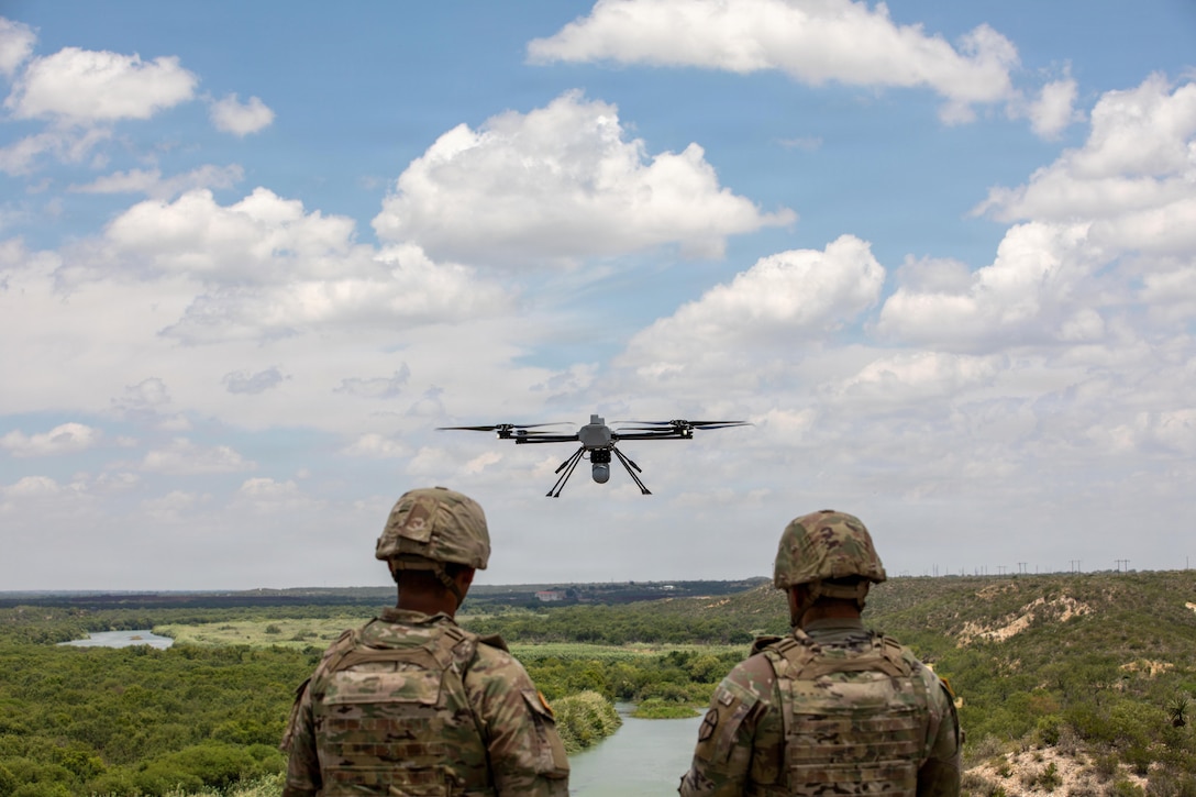 Two soldiers, shown from behind, face toward a drone hovering in the near distance above a field.