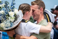 A sailor in a white uniform hugs their spouse as they hold blue and white flowers.