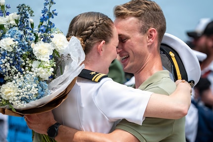 A sailor in a white uniform hugs their spouse as they hold blue and white flowers.