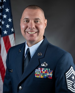 West Virginia Air National Guard Command Chief Master Sgt. Scott Melton official portrait. Man smiling in Air Force dress blue uniform in front of draped American Flag.