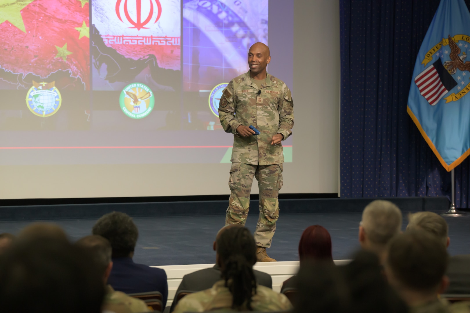 Air Force Chief Master Sgt. Kenneth Bruce speaks onstage with emblems for the US Pacific Command, US Central Command, and War Department accompained by graphics featuring the Chinese flag, Iranian flag and US dollar.