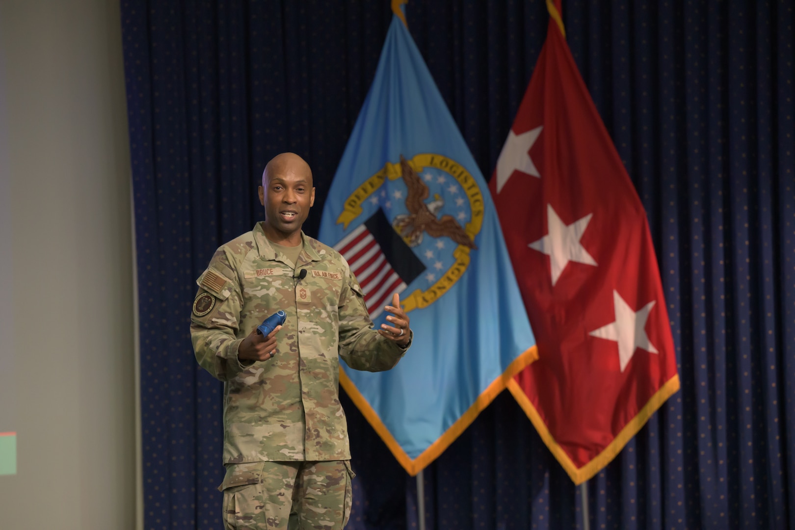 Air Force Chief Master Sgt. Kenneth Bruce speaks on a stage in front of a DLA flag and a three-star flag