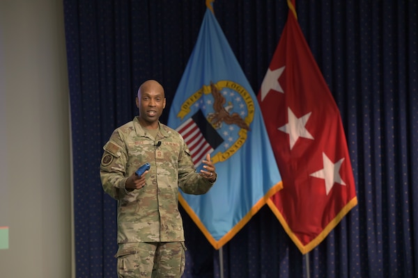 Air Force Chief Master Sgt. Kenneth Bruce speaks on a stage in front of a DLA flag and a three-star flag