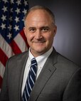A smiling person in business attire poses for a professional photo in front of the U.S. flag in a portrait studio.