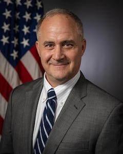 A smiling person in business attire poses for a professional photo in front of the U.S. flag in a portrait studio.