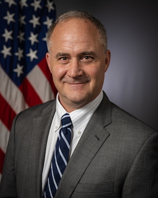A smiling person in business attire poses for a professional photo in front of the U.S. flag in a portrait studio.