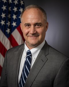 A smiling person in business attire poses for a professional photo in front of the U.S. flag in a portrait studio.