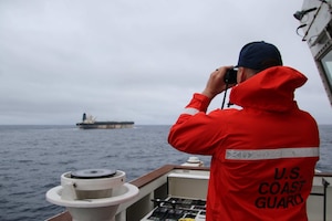 A man wearing an orange jacket looks through binoculars to monitor a ship sailing along the ocean; the back of the jacket reads "U.S. Coast Guard."