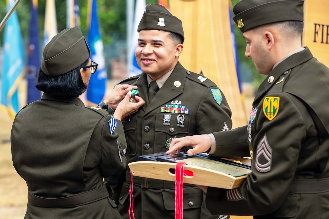 A soldier pins a medal to a smiling soldier's lapel as another soldier holding a tray with plaques stands to the right.