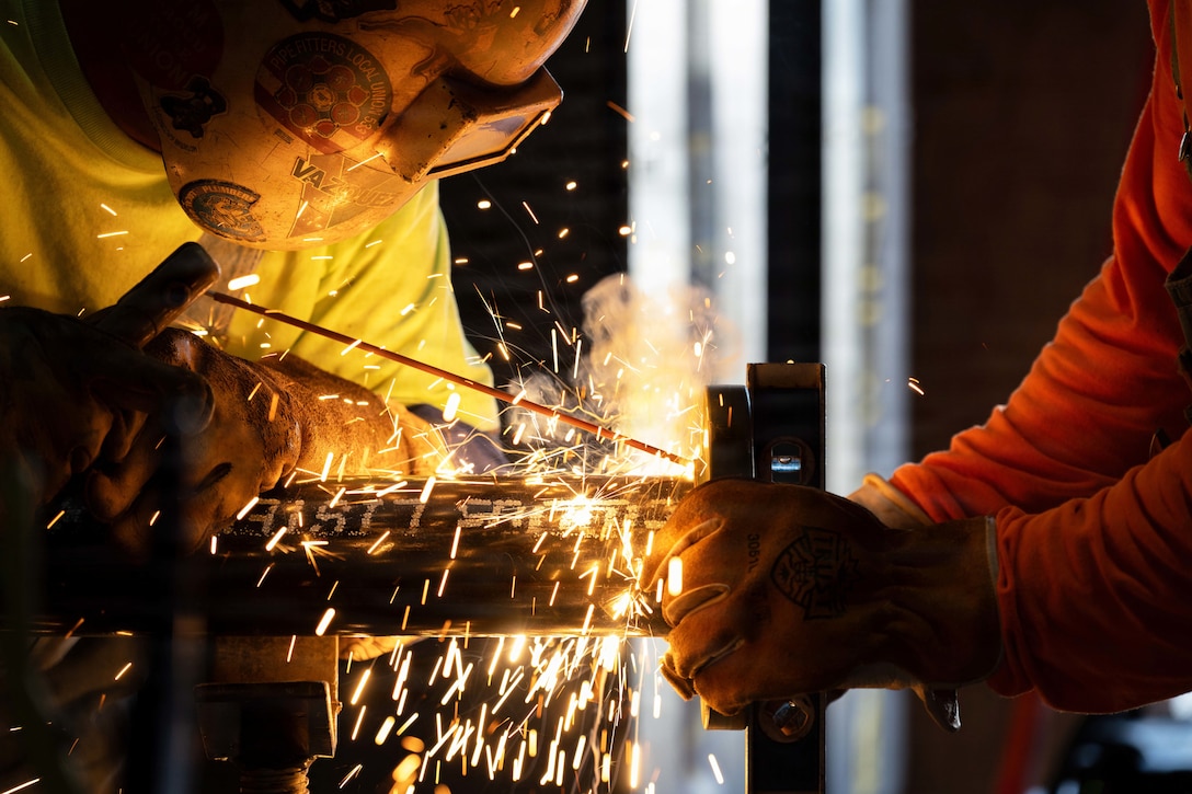 Sparks fly as a contractor wearing protective gear welds a pipe in a dark room.