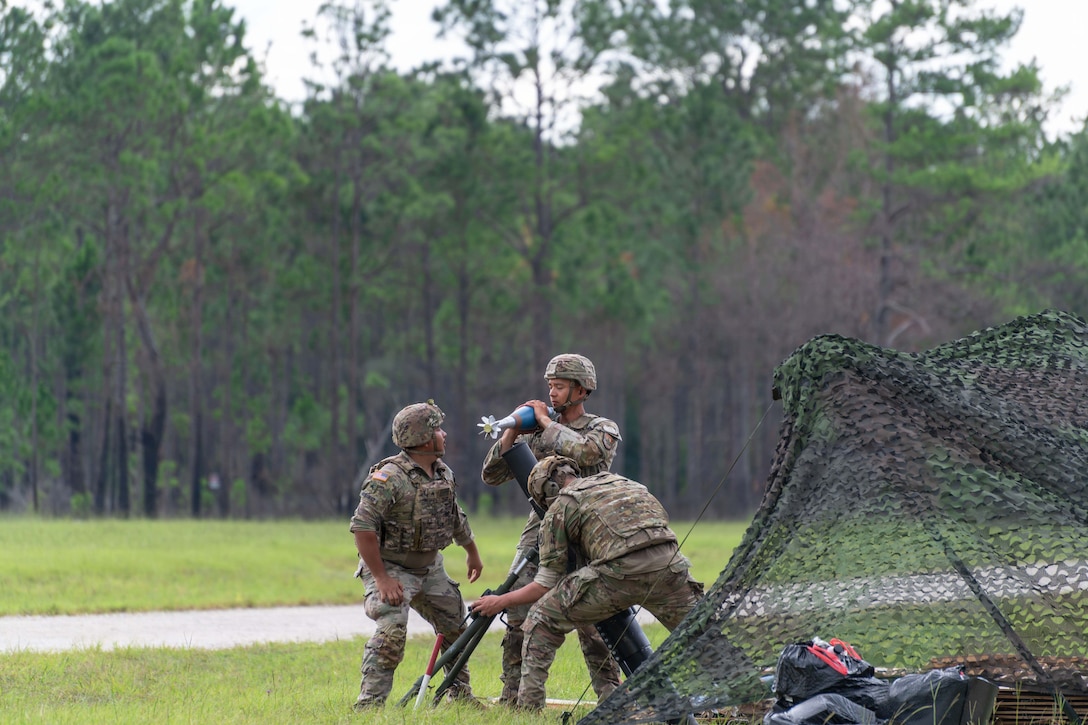 Three soldiers in camouflage uniforms load a weapon in two a tripod system near a netted tent in a wooded area.