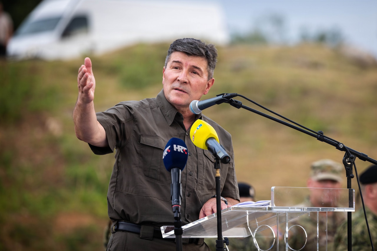 A man stands at a lectern and points to his right.