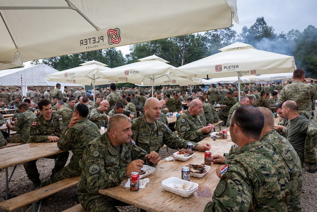 Groups of people wearing camouflage military uniforms sit at tables under canopies for a meal.
