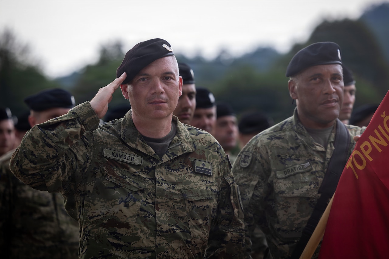 A man wearing a camouflage military uniform renders a salute while holding a flag. Other people in similar attire stand in formation.