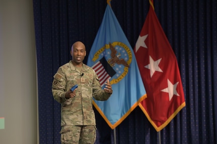Air Force Chief Master Sgt. Kenneth Bruce speaks on a stage in front of a DLA flag and a three-star flag