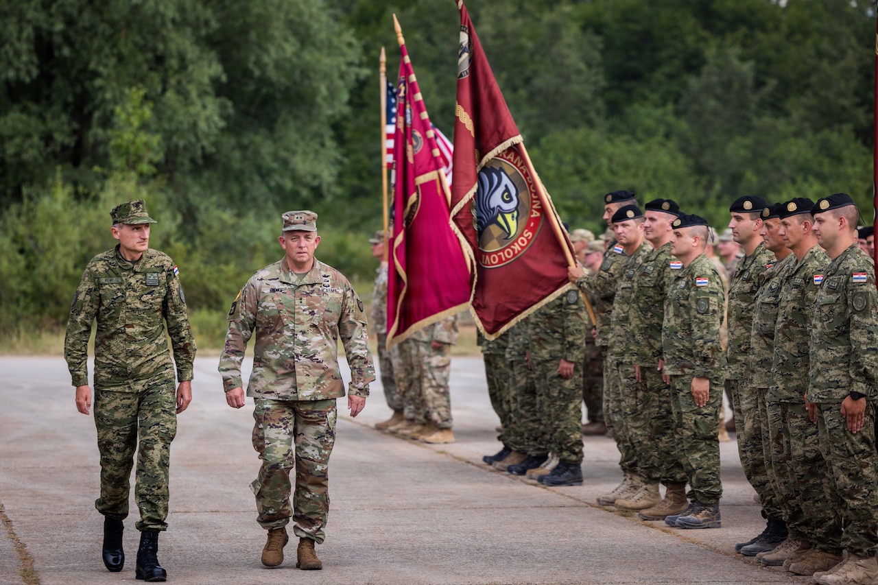 Two men wearing camouflage military uniforms walk past a formation of military personnel holding various flags.