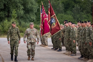 Two men wearing camouflage military uniforms walk past a formation of military personnel holding various flags.