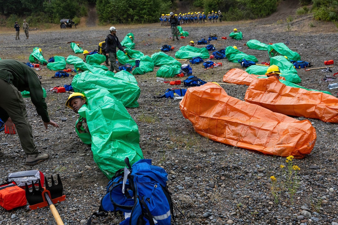 People lie on the ground outdoors in green and orange plastic bags with their heads exposed as a few people stand and give instructions.