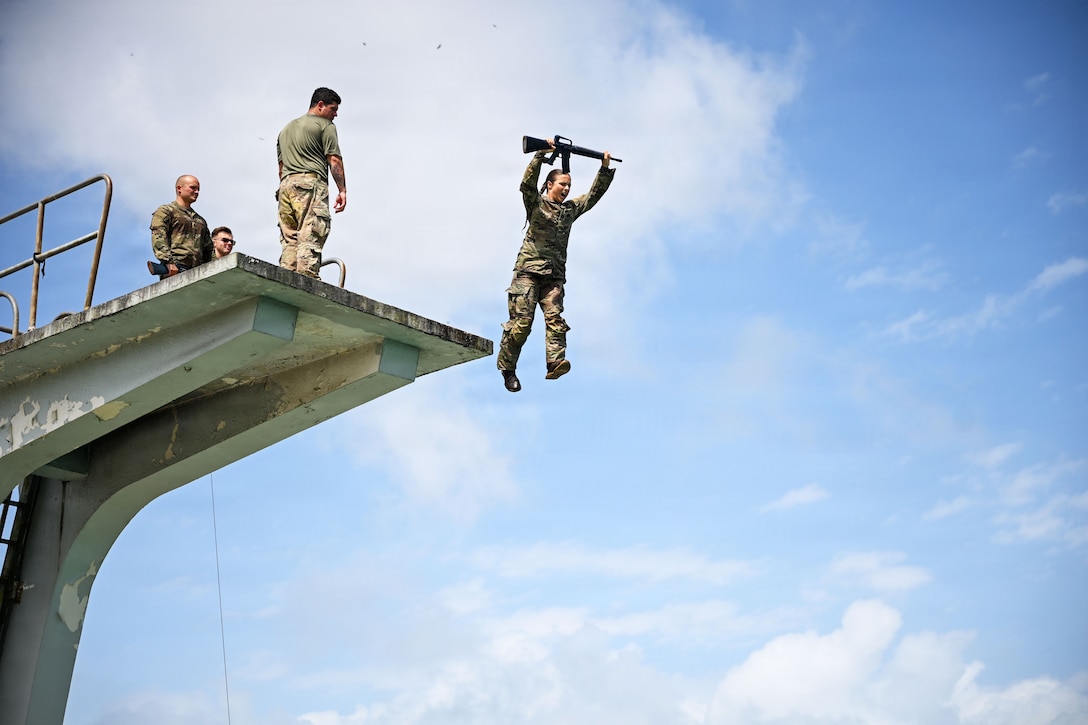 A soldier holding a rifle in the air jumps off a high diving platform. Other soldiers standing on the platform watch.