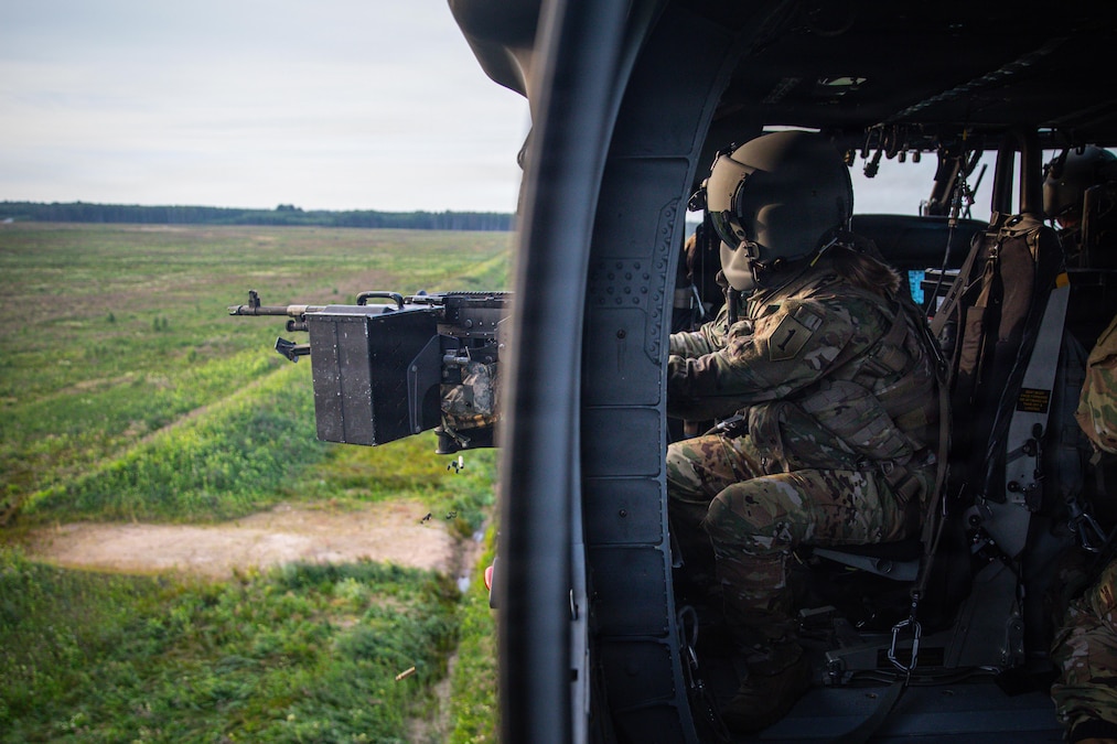 A soldier in a helicopter that is low to the ground over a green field looks out the window while holding onto a machine gun positioned over the field.