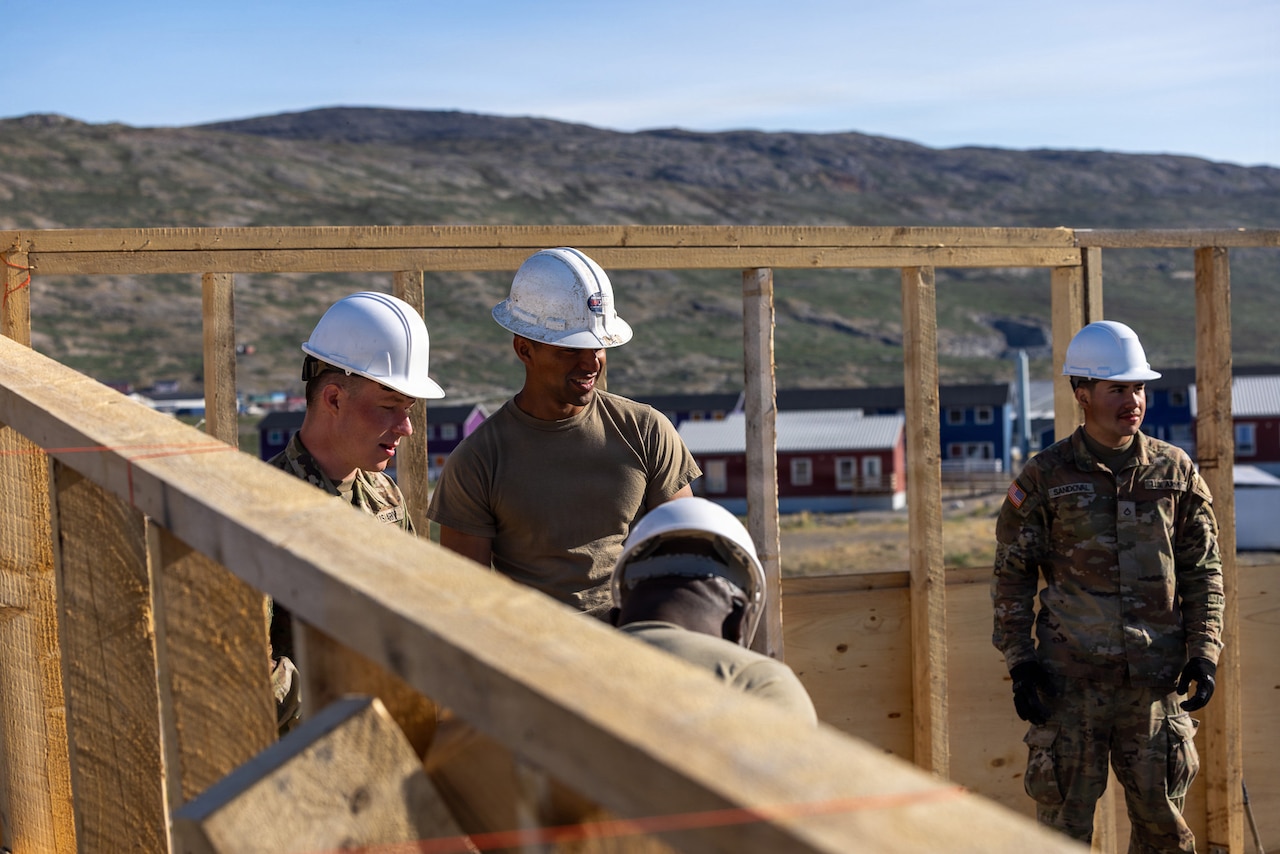 Four people wearing camouflage military uniforms and hard hats stand outside in a wooden building frame.