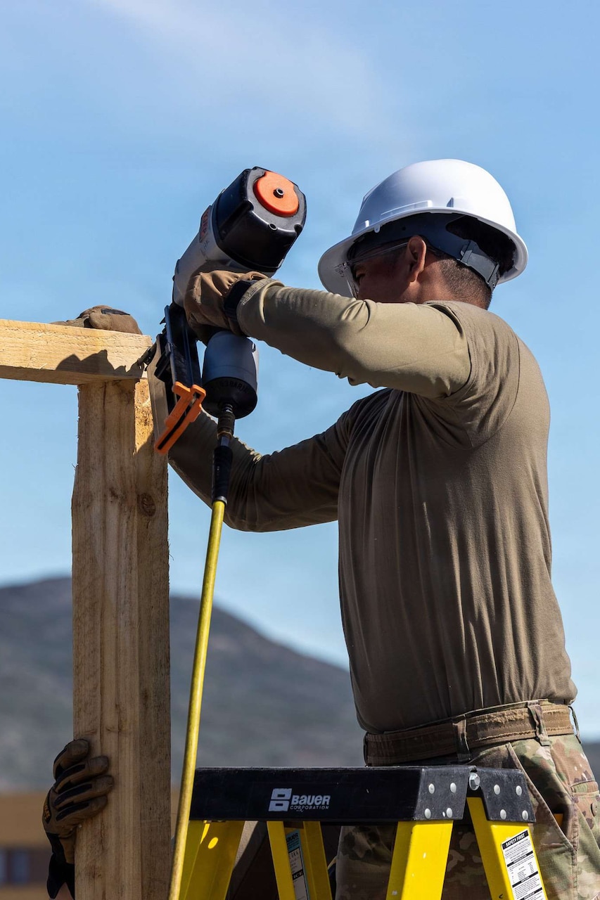 A man wearing a camouflage military uniform and hard hat stands on a ladder and uses a power tool to secure two wooden beams.