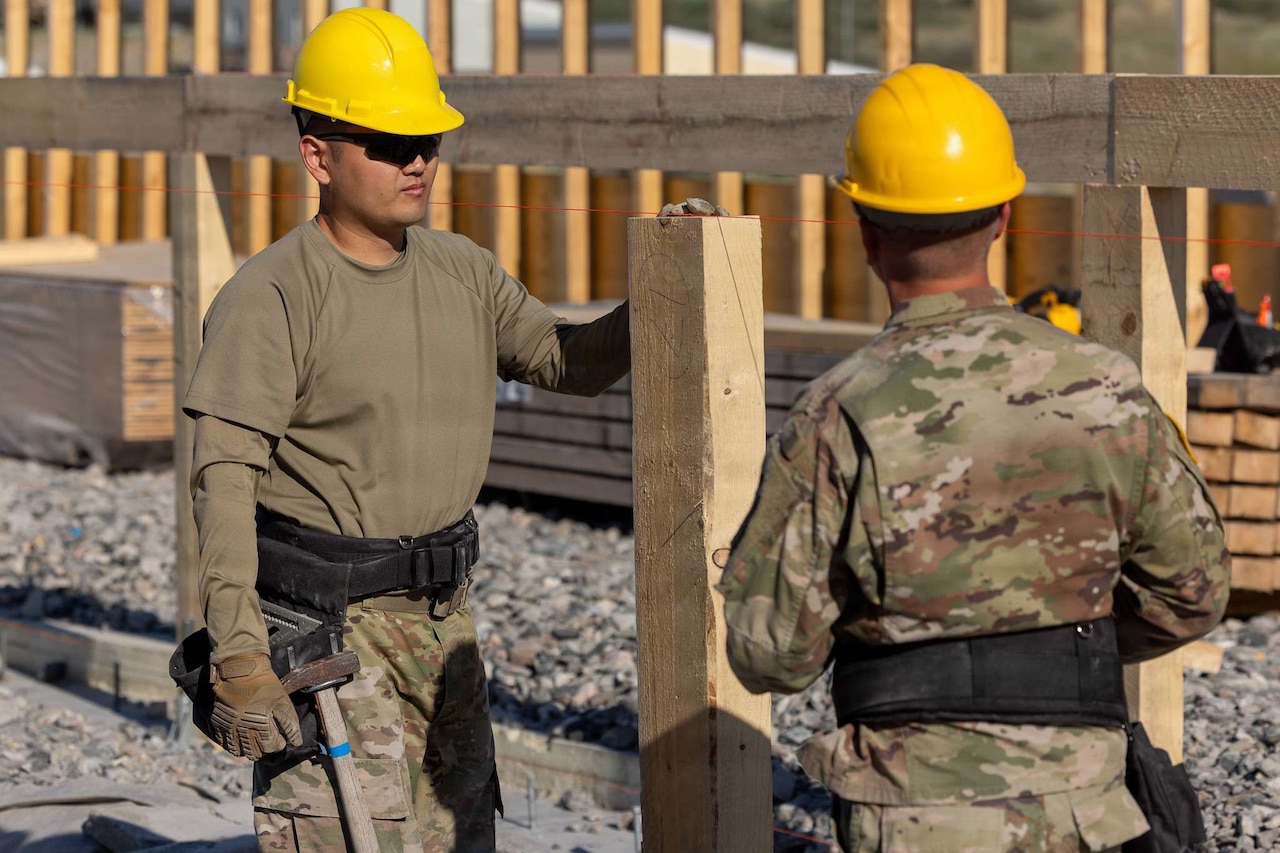 Two men wearing camouflage military uniforms and hard hats stand outside holding power tools next to a wooden building frame.