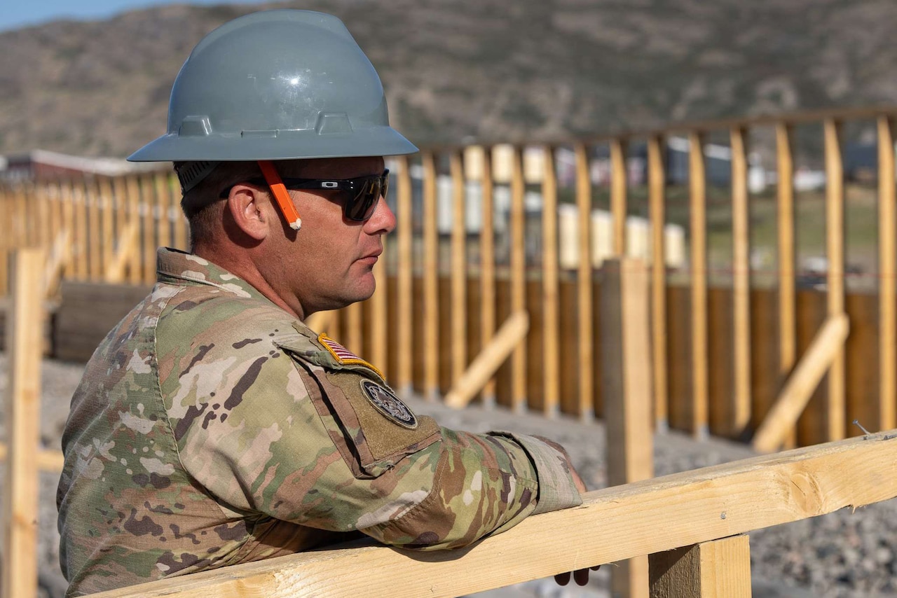 A man wearing a camouflage military uniform, hard hat and sunglasses leans against a wooden rail outside and watches something out of frame.