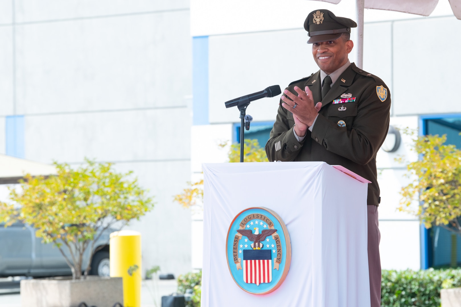 A man wearing a military uniform stands behind a podium, outside on a stage under a tent, speaking to a crowd.