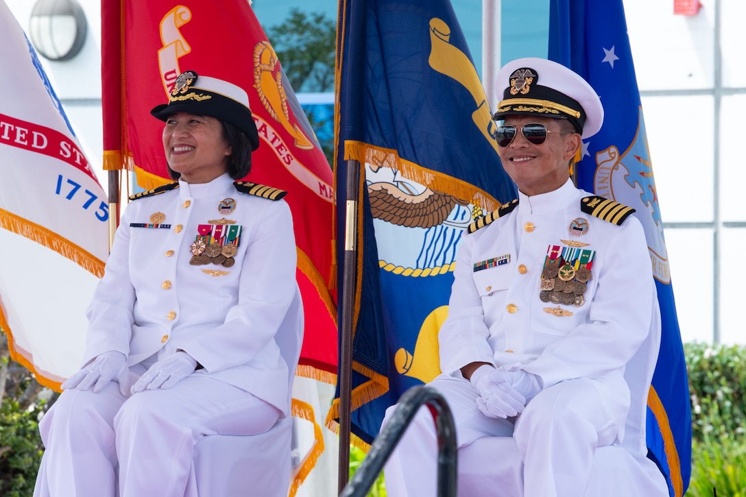A woman and a man, both wearing military uniforms, sit outside on a stage covered by a tent, smiling.