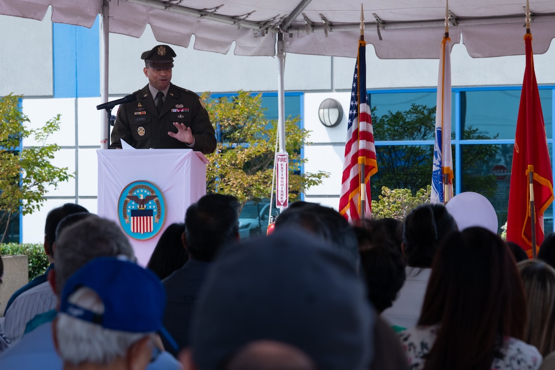 A man wearing a military uniform stands behind a podium, outside on a stage under a tent, speaking to a crowd.