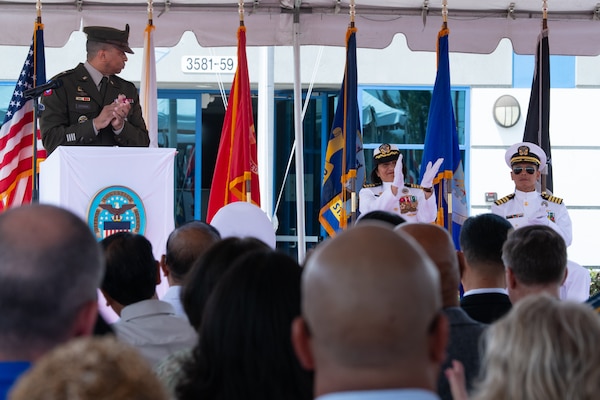 A man wearing a military uniform stands behind a podium, outside on a stage under a tent, speaking to a crowd.