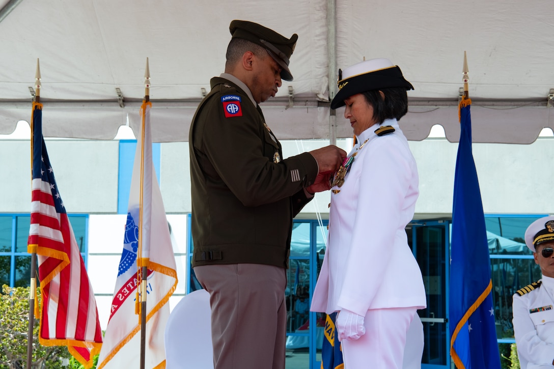 A man wearing a military uniform stands outside on a stage covered by a tent pinning a medal on a woman wearing a military uniform.