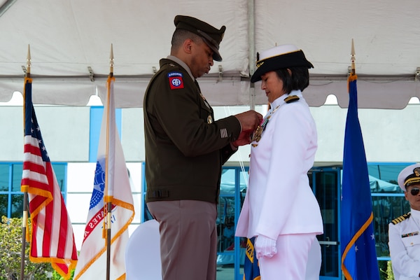 A man wearing a military uniform stands outside on a stage covered by a tent pinning a medal on a woman wearing a military uniform.