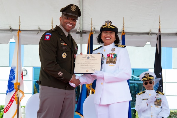 A man and woman wearing military uniforms stand outside on a stage covered by a tent holding a certificate