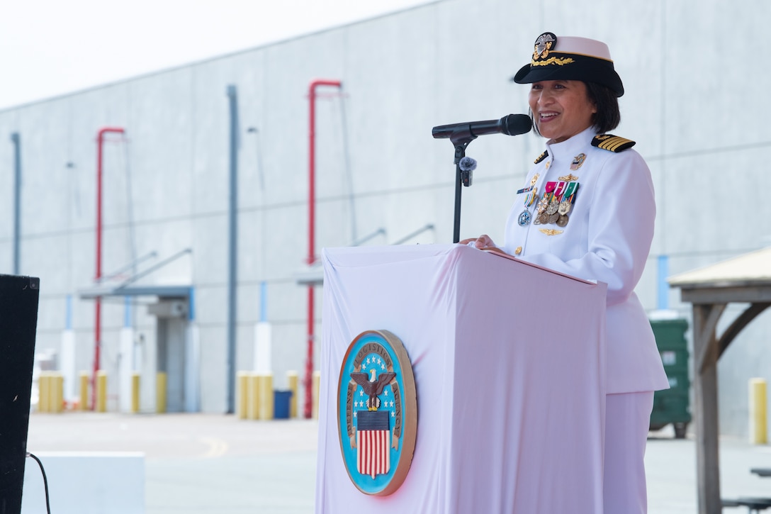 A woman wearing a military uniform stands behind a podium, outside on a stage under a tent, speaking to a crowd.